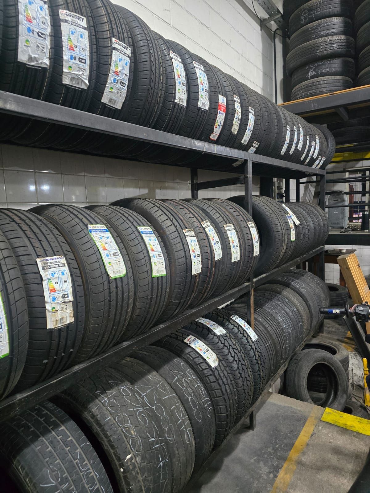 Neumáticos apilados en estantes de metal dentro de una tienda, mostrando varios patrones de banda de rodadura y etiquetas de eficiencia energética.