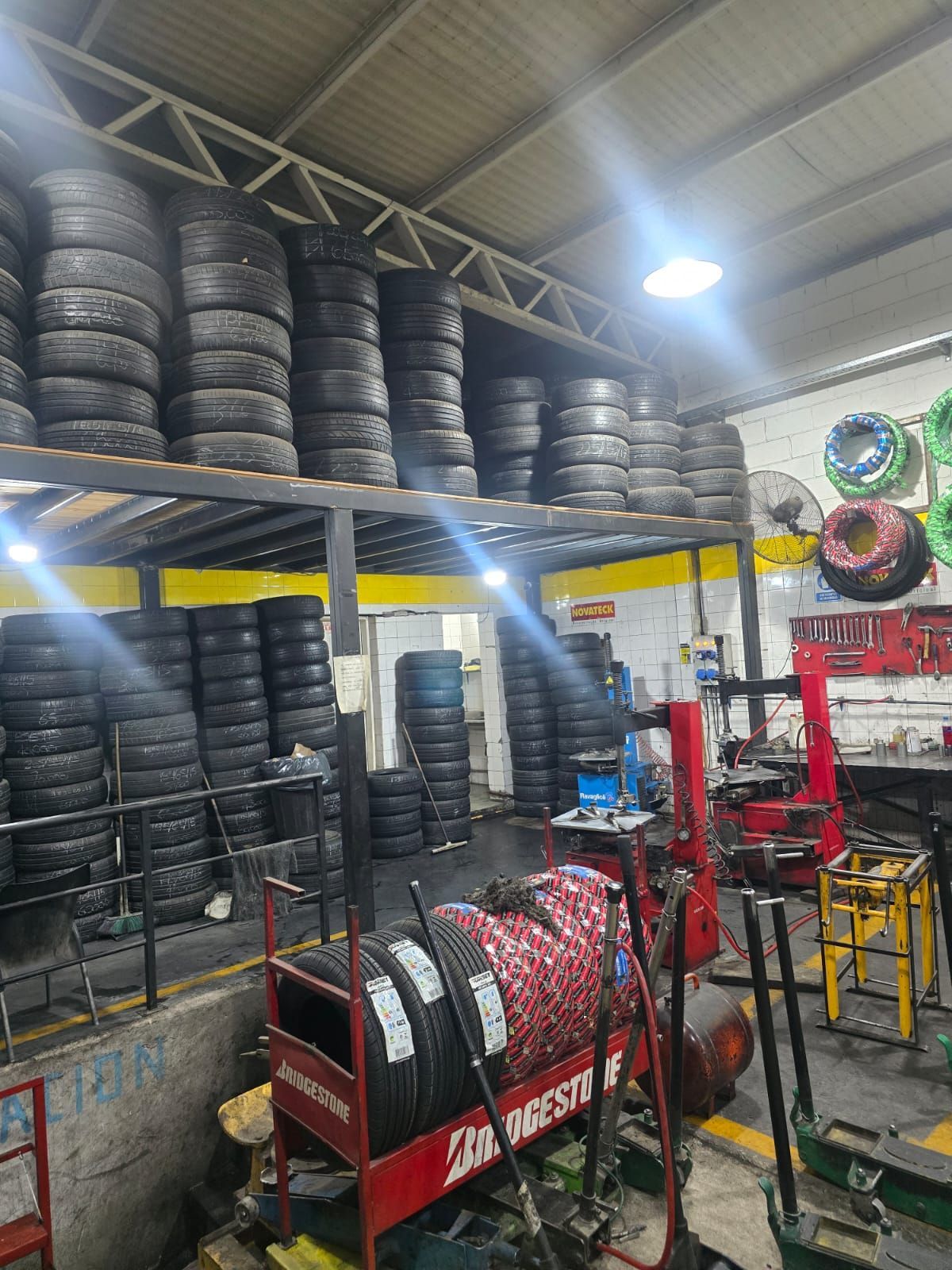 Interior de una tienda de neumáticos con pilas de neumáticos en estanterías. También se ven neumáticos Bridgestone rojos en un carrito, maquinaria y herramientas.