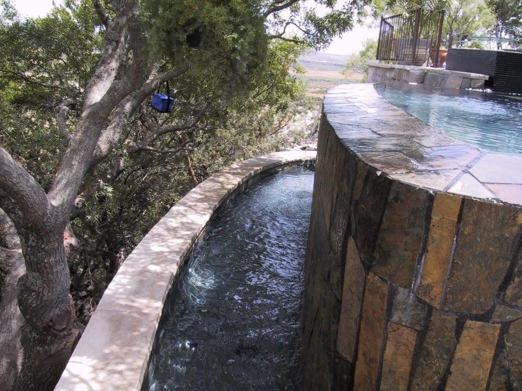 Water cascading from stone wall into a narrow, stone-lined pool, with a tree in the background.