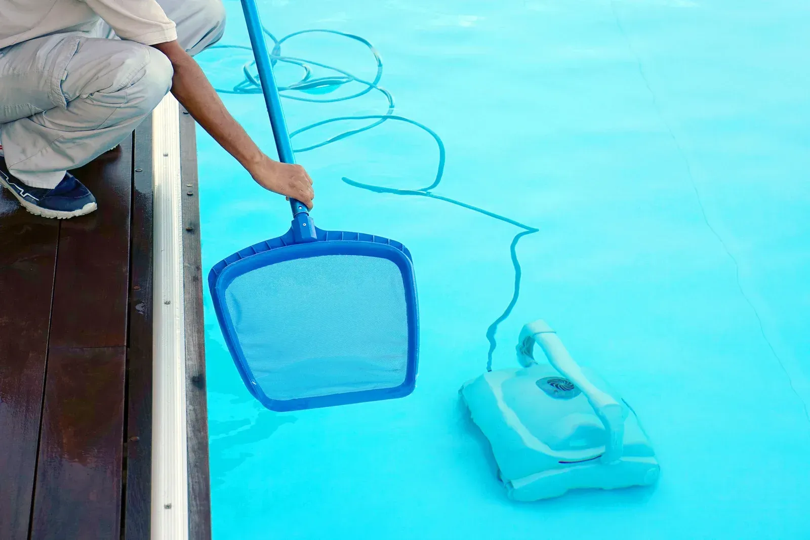 Person using a net to collect debris near a blue robotic pool cleaner in a turquoise pool.