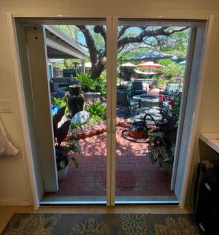 A white retractable screen door frame overlooking a brick patio with lush plants, patio furniture, and a large tree.