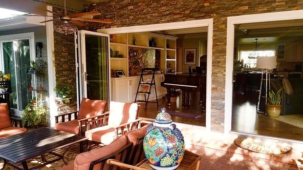 A sunny patio with metal chairs and a table leads into an indoor living space featuring a piano and stone accent wall.