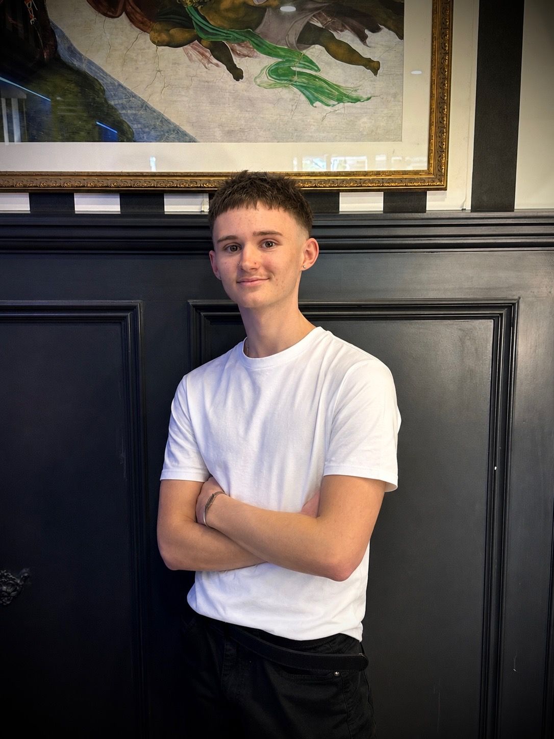 Person in a white T-shirt standing with arms crossed in front of a dark paneled wall. — Connolly & Co Barbers In Robina, QLD
