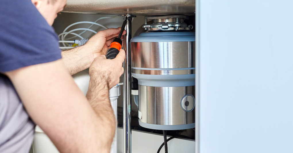 Person working on a kitchen garbage disposal with a screwdriver under a sink.