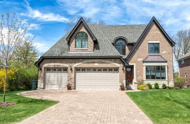 Suburban brick house with two garages, steep roof, and a paved driveway under a blue sky