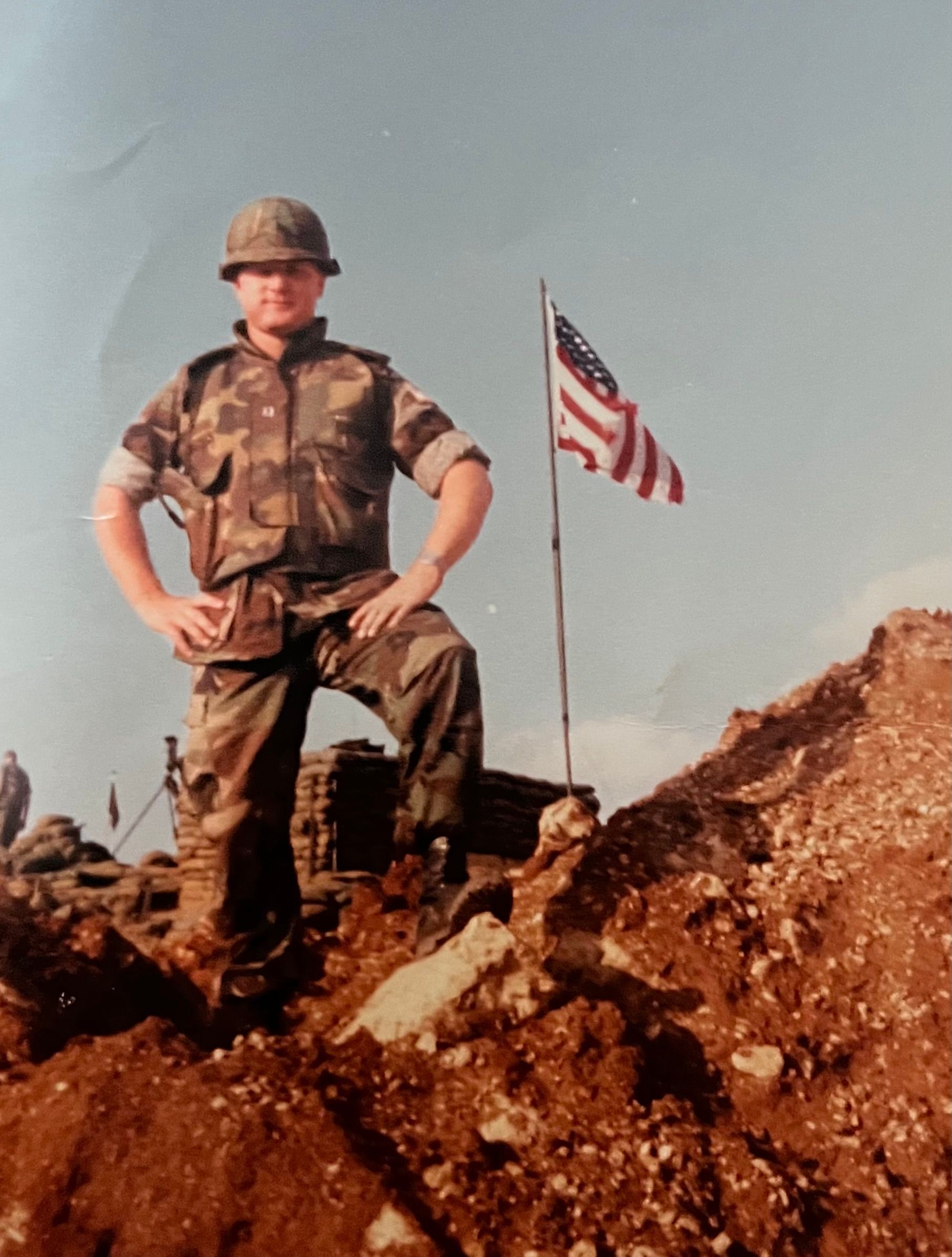 A soldier stands on top of a pile of dirt in front of an american flag