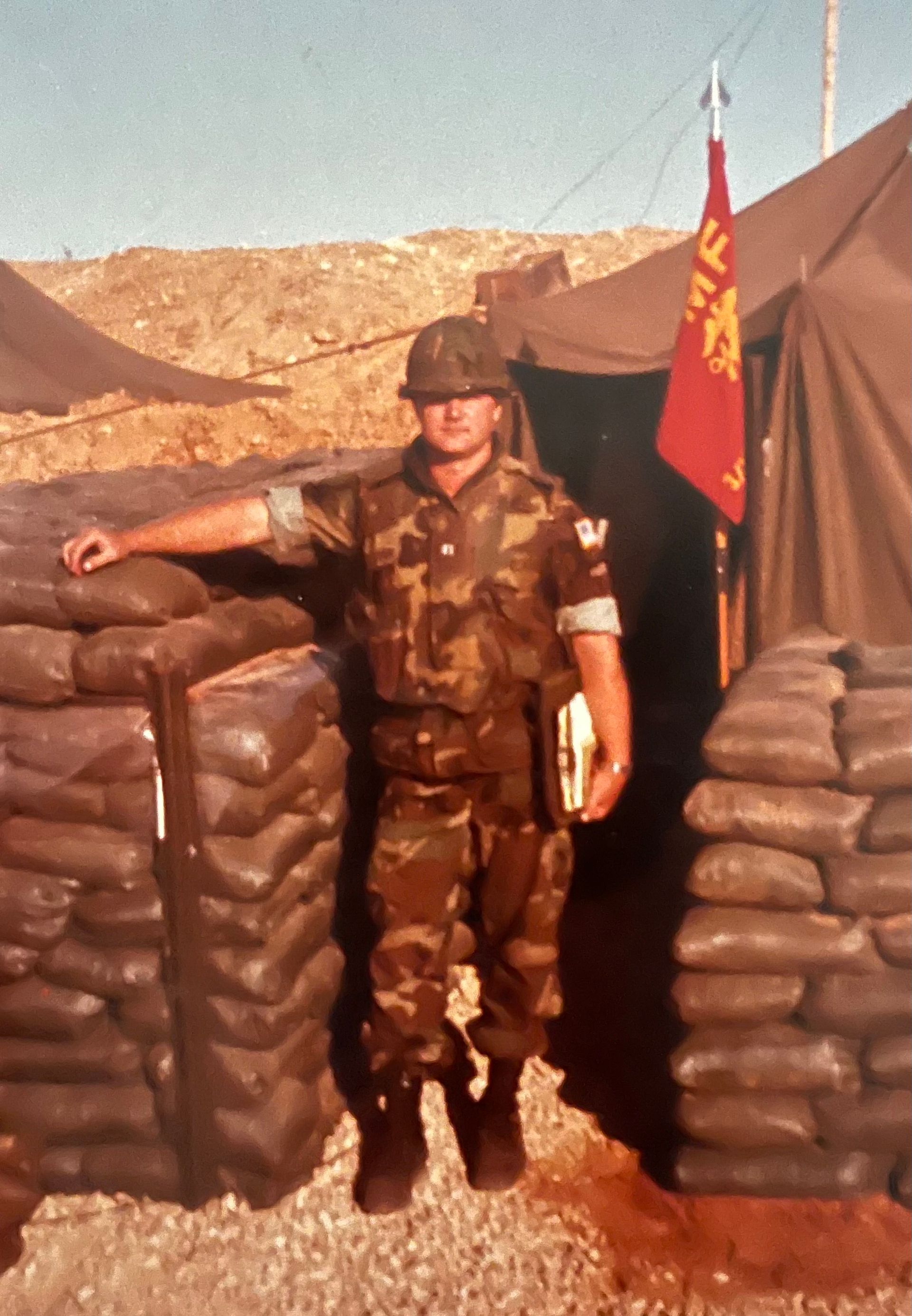 A man in a military uniform stands next to a pile of sandbags