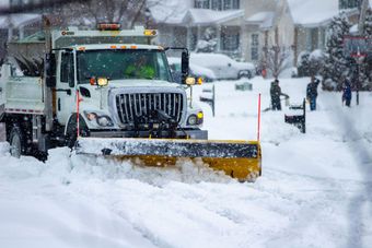 Front view of city services snow plow truck with yellow blade cleaning roads after winter storm with kids playing in the background