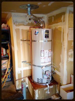 A water heater installed in a corner. It has a vent pipe, copper pipes, and sits on a wooden base.