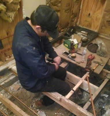 Person in a baseball cap works on plumbing in a partially demolished room. Copper pipes and tools are scattered.
