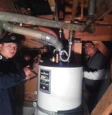 Two people stand near a new water heater in a dimly lit basement. Plumbing pipes are overhead.