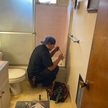 Plumber installing a bathroom fixture in a pink-tiled space. He's kneeling and using tools from a toolbox on the floor.
