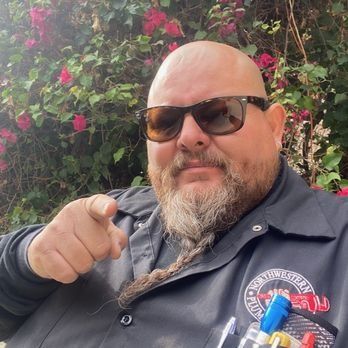 Man with sunglasses and beard points at viewer, standing outside near pink flowers.