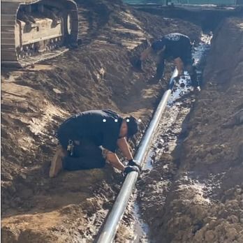 Two workers installing a metal pipe in a trench, next to an excavator.