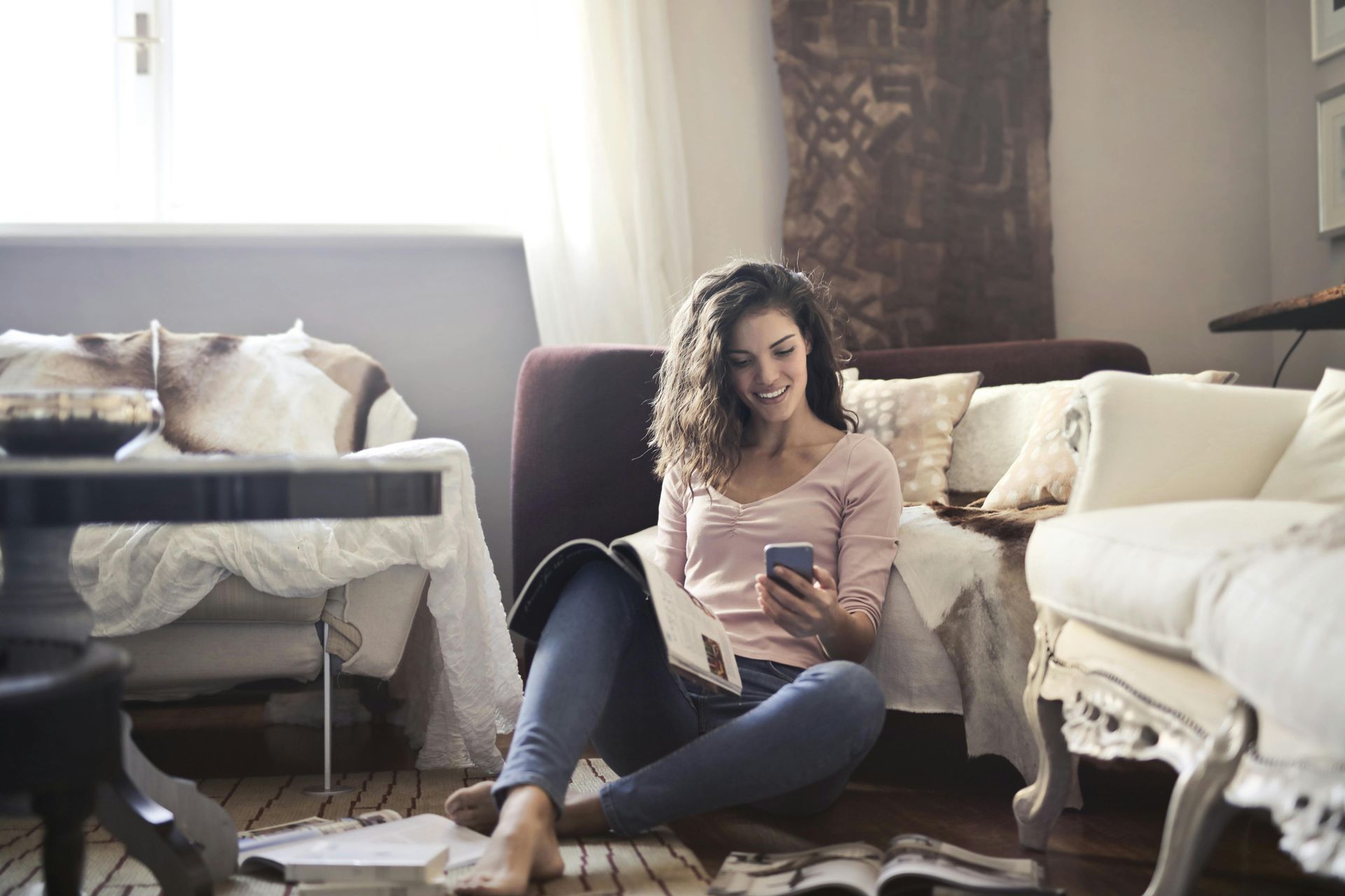 A person sits on a rug in a bright, cozy living room, smiling while looking at a smartphone and holding an open magazine.
