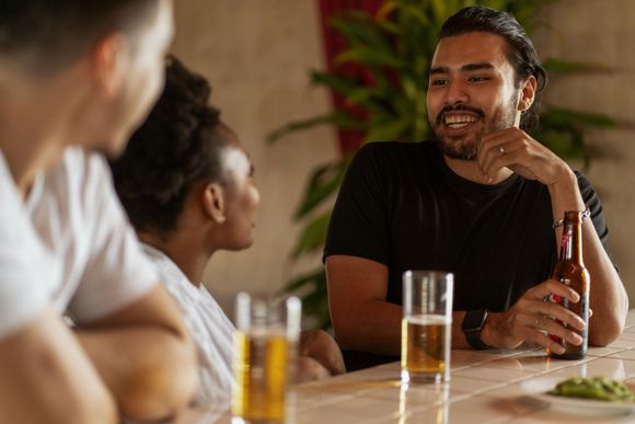Three people socialize at a bar with beer and guacamole, one individual smiling while holding a bottle.