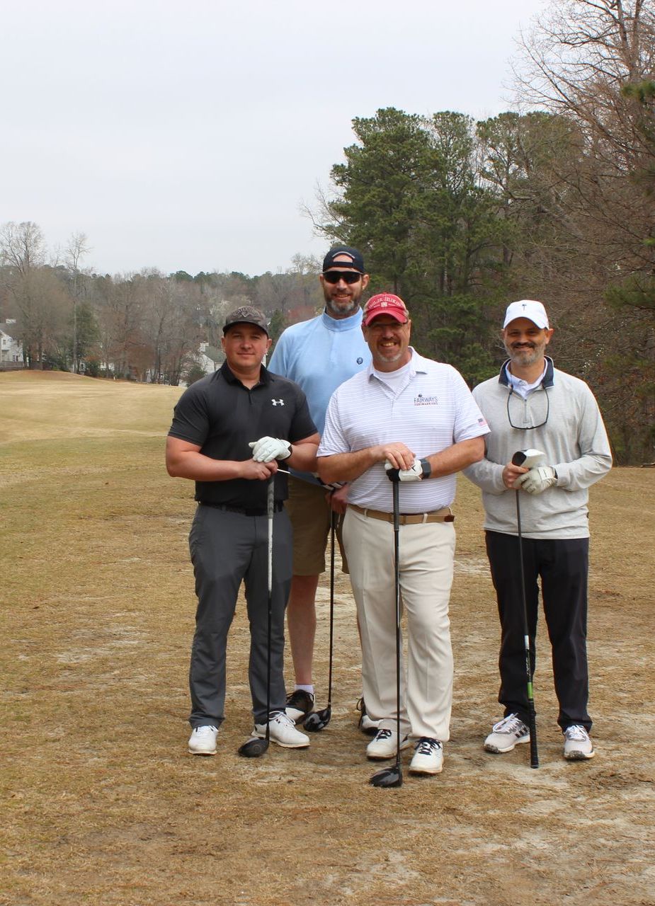 Four men pose on a golf course, holding golf clubs. The men are smiling and looking at the camera.