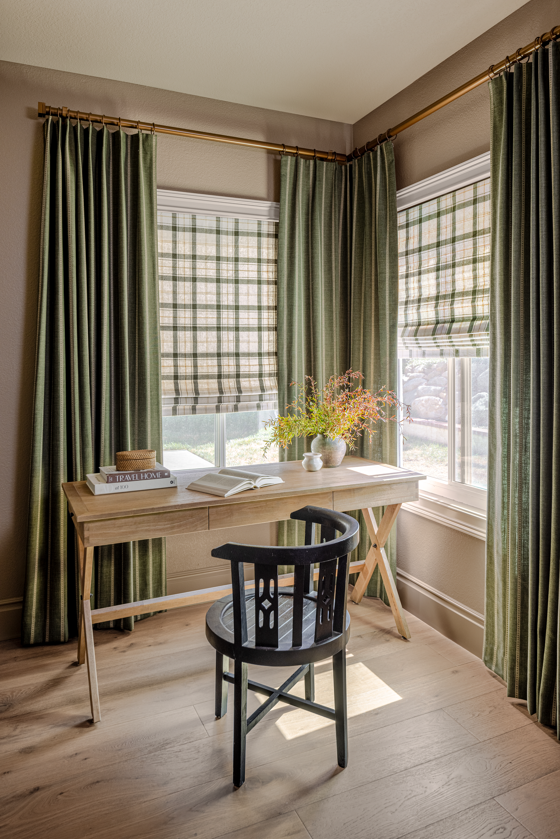 A bright, rustic home office with a wooden desk, black chair, and green curtains framing the windows.
