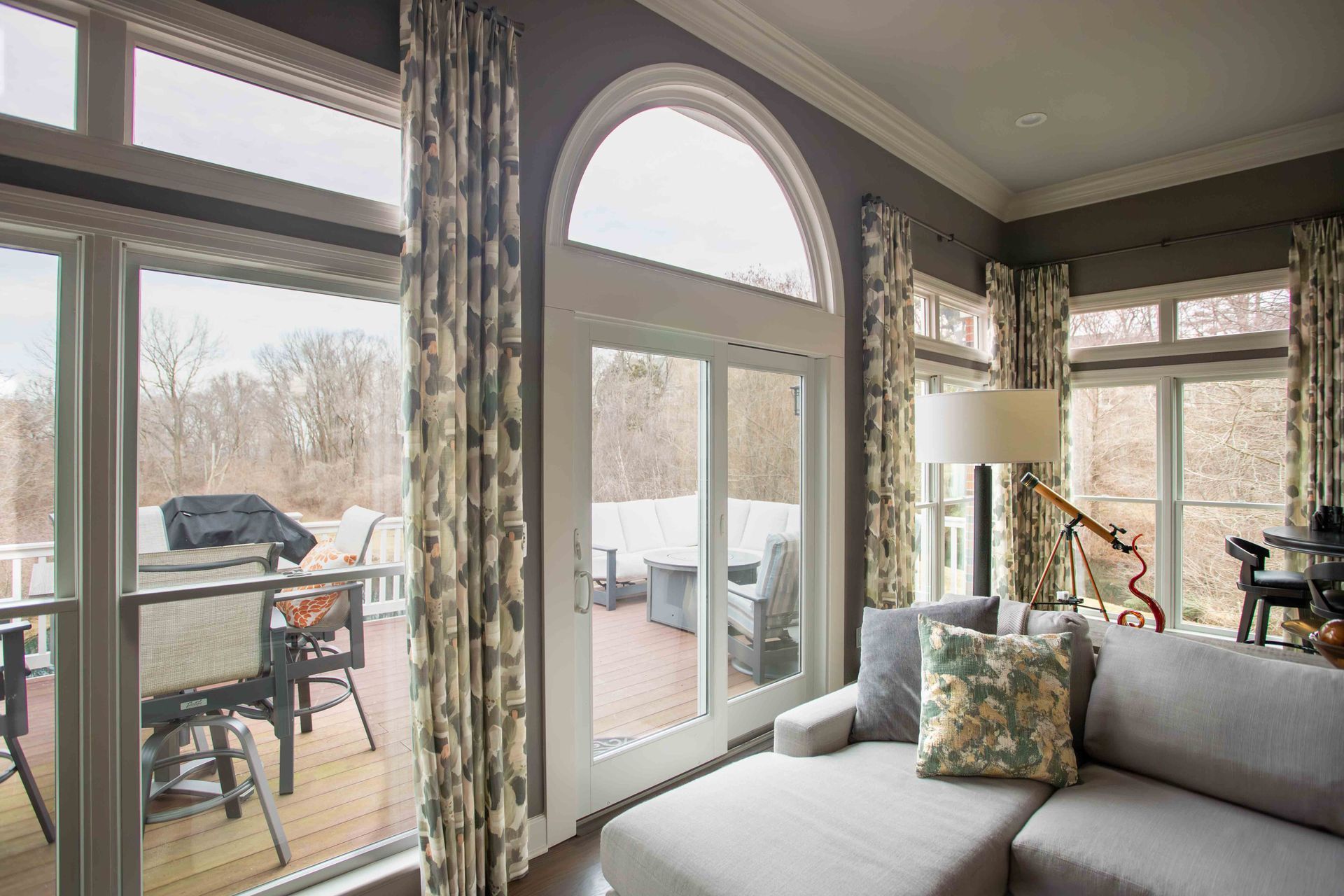 Living room with large windows overlooking a deck and trees. Gray walls, curtains, and a gray sofa.