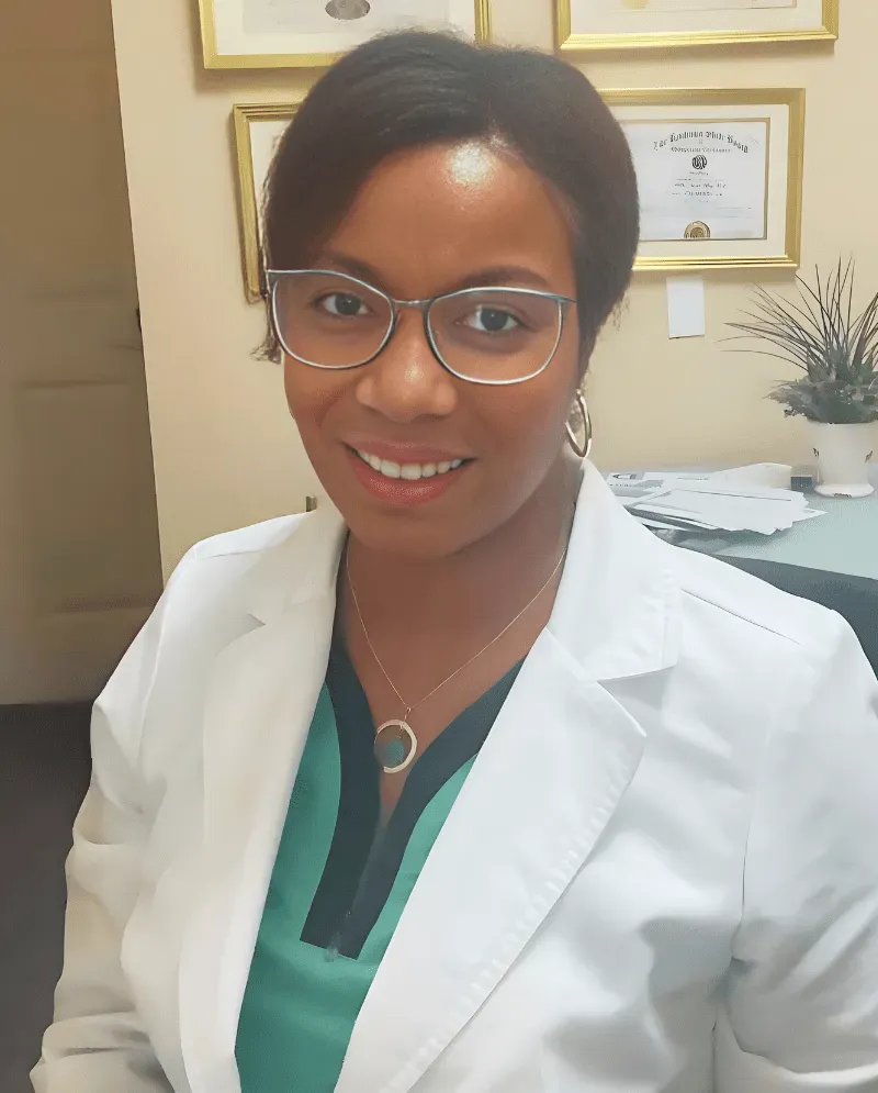 Woman wearing glasses and a white coat, smiling, in an office setting.