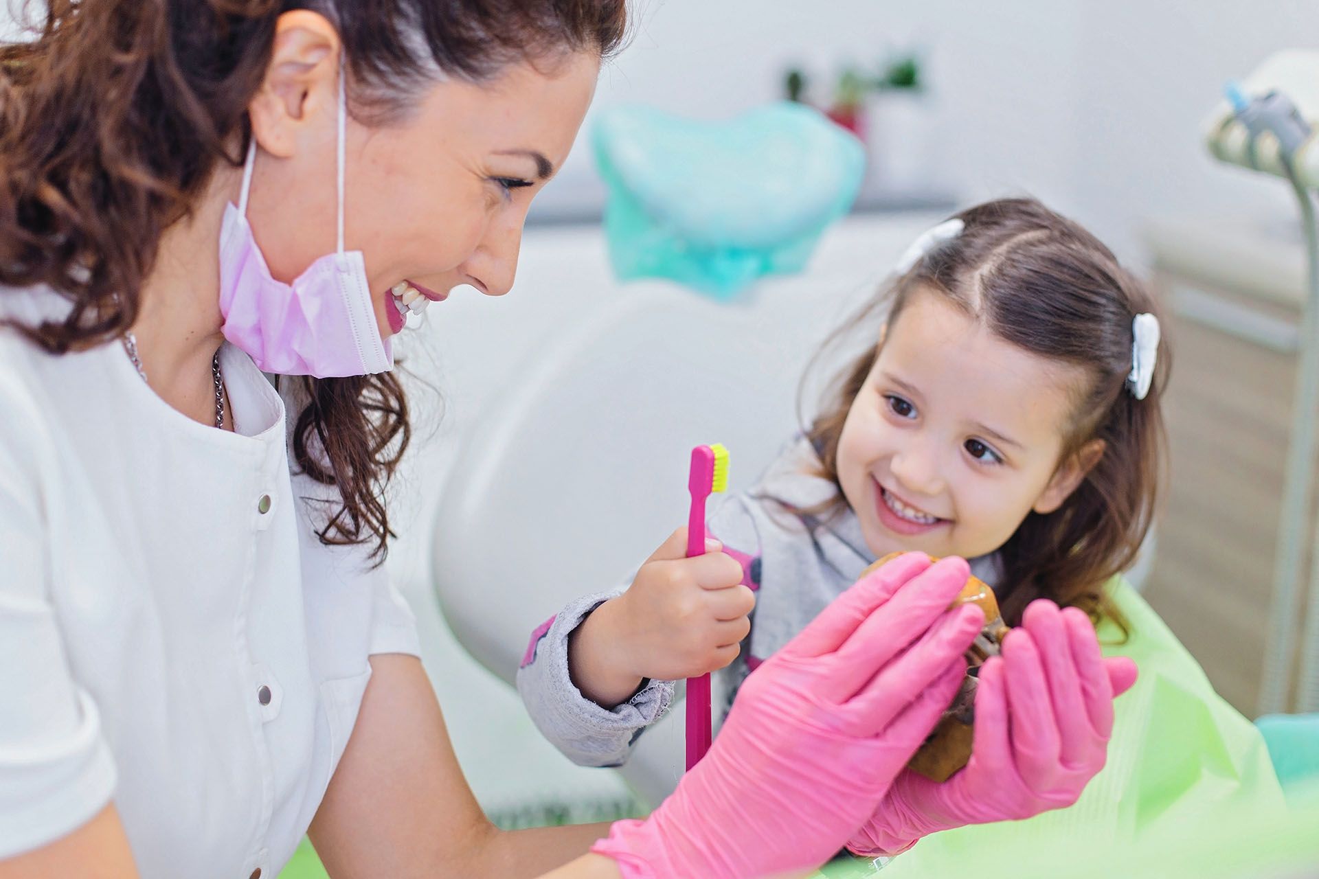 Dentist shows a child how to brush toy teeth with pink toothbrush in a dental office.
