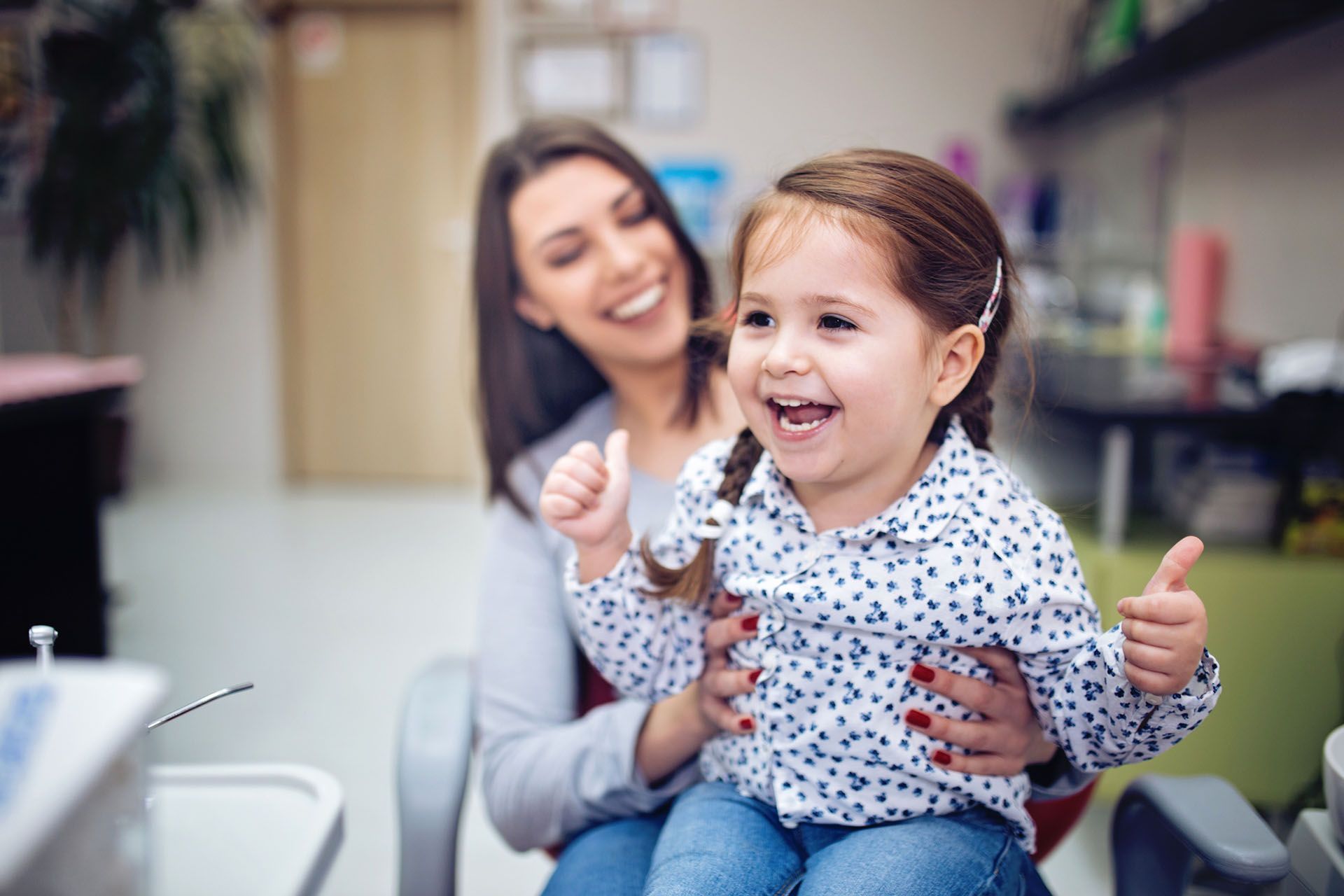 Mother holding smiling child at a dentist's office, both giving thumbs up.
