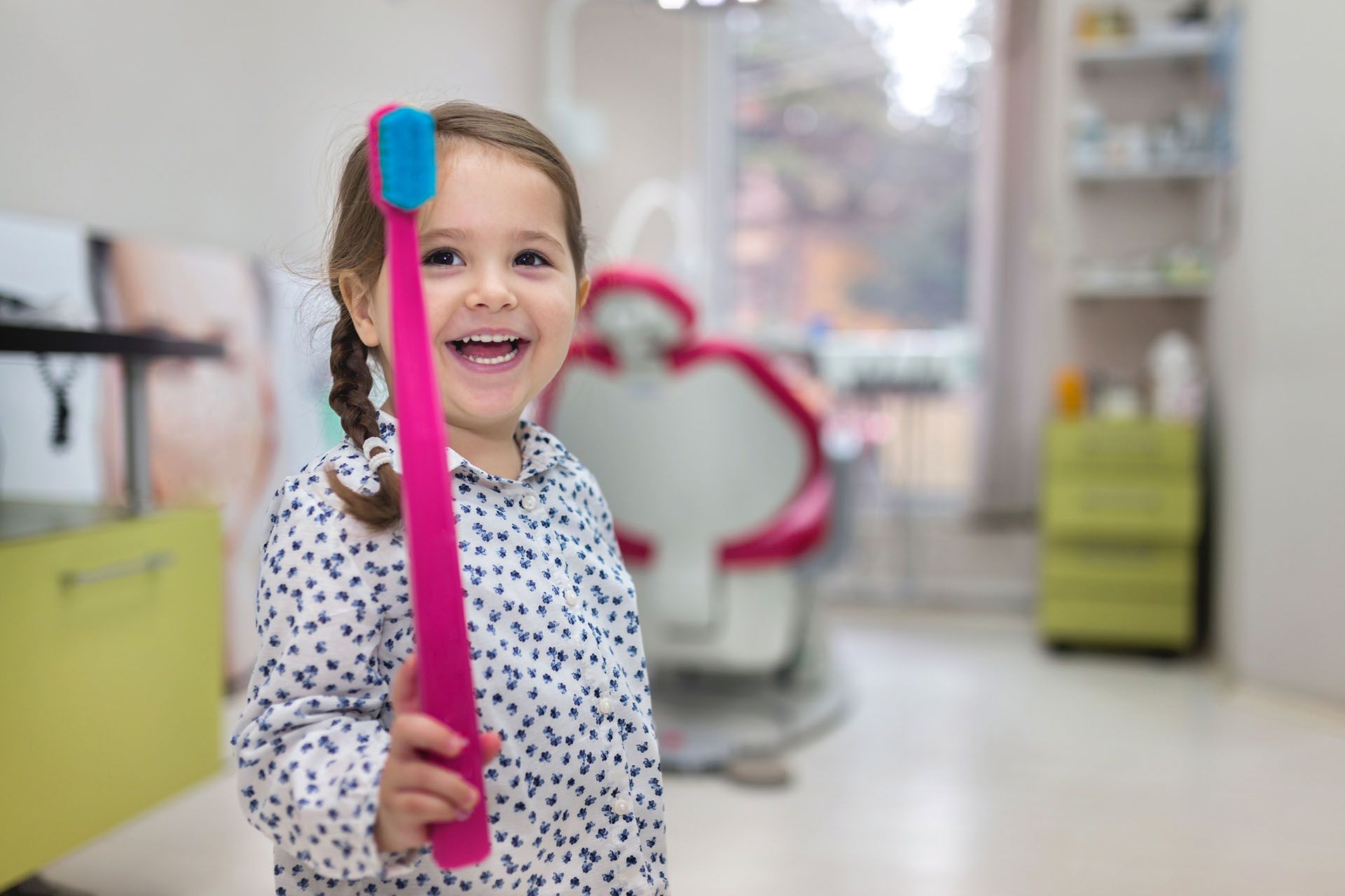 Girl smiling, holding giant pink toothbrush in a dentist's office.