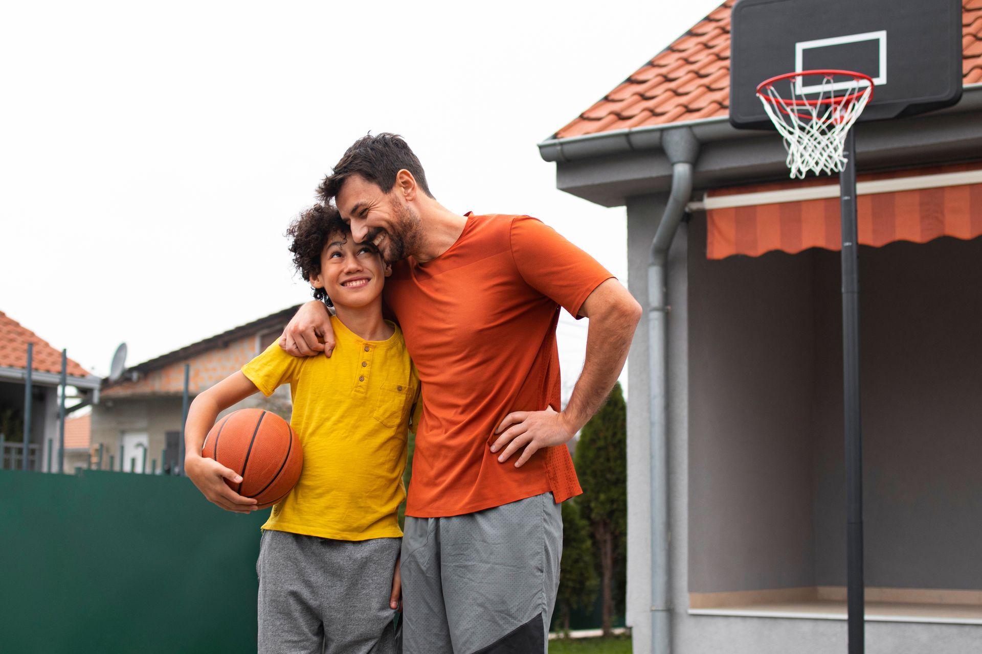 Man with arm around a boy holding a basketball, standing near a hoop in front of a house.