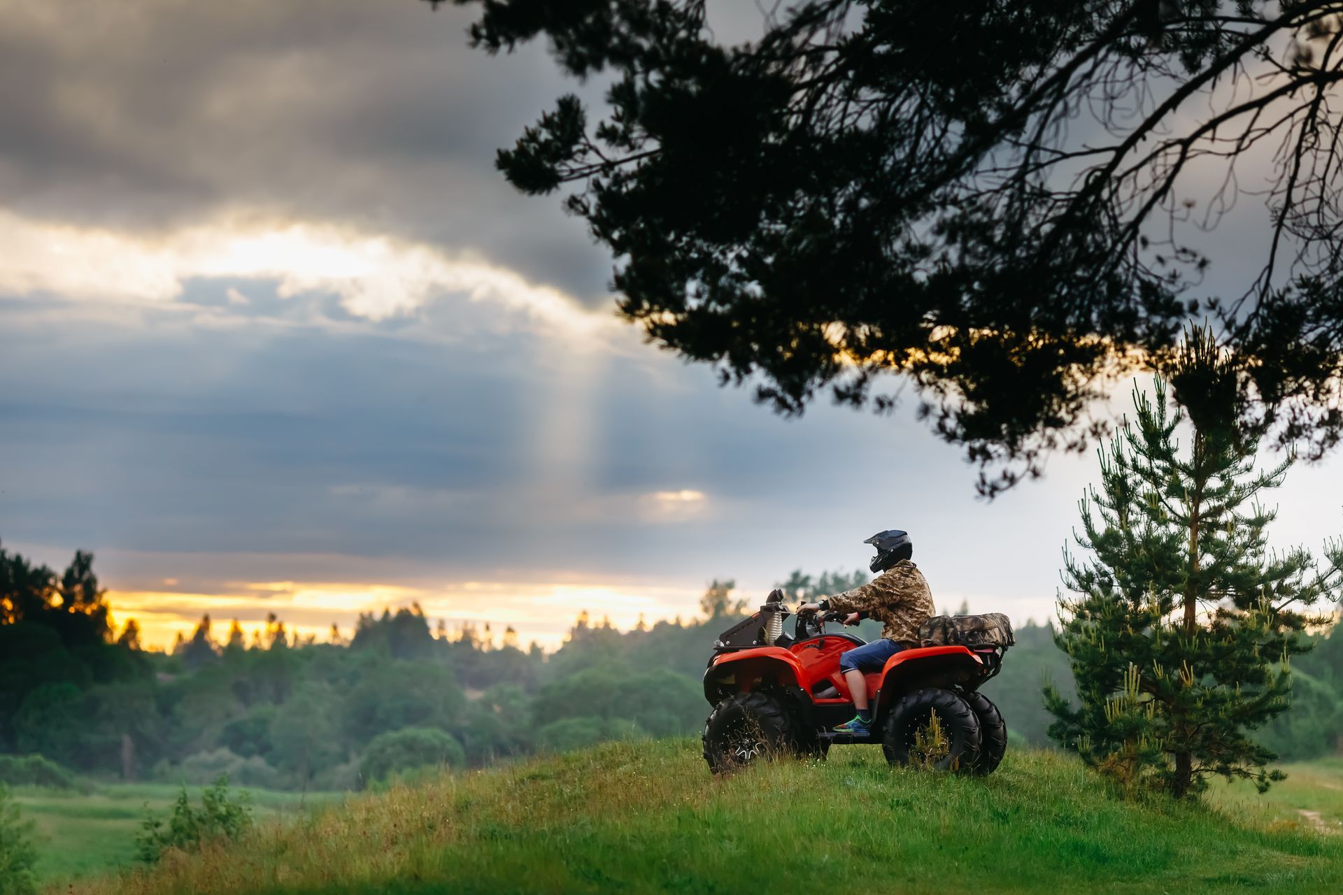 Person riding a red ATV on a grassy hill, with a forest in the background and a cloudy sky at sunset.