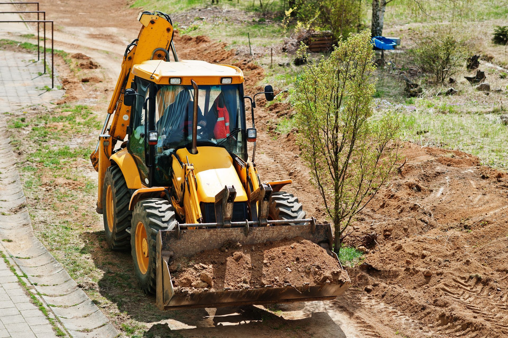 Yellow backhoe moving dirt on a construction site next to a sidewalk and small tree.
