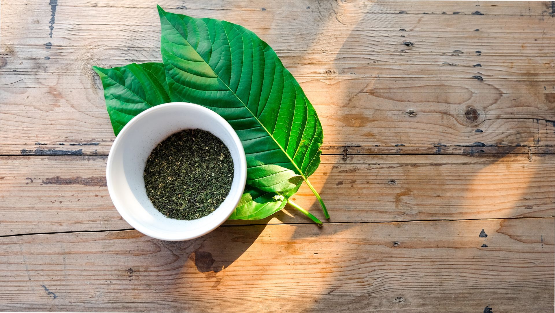 A bowl of green tea next to a green leaf on a wooden table.