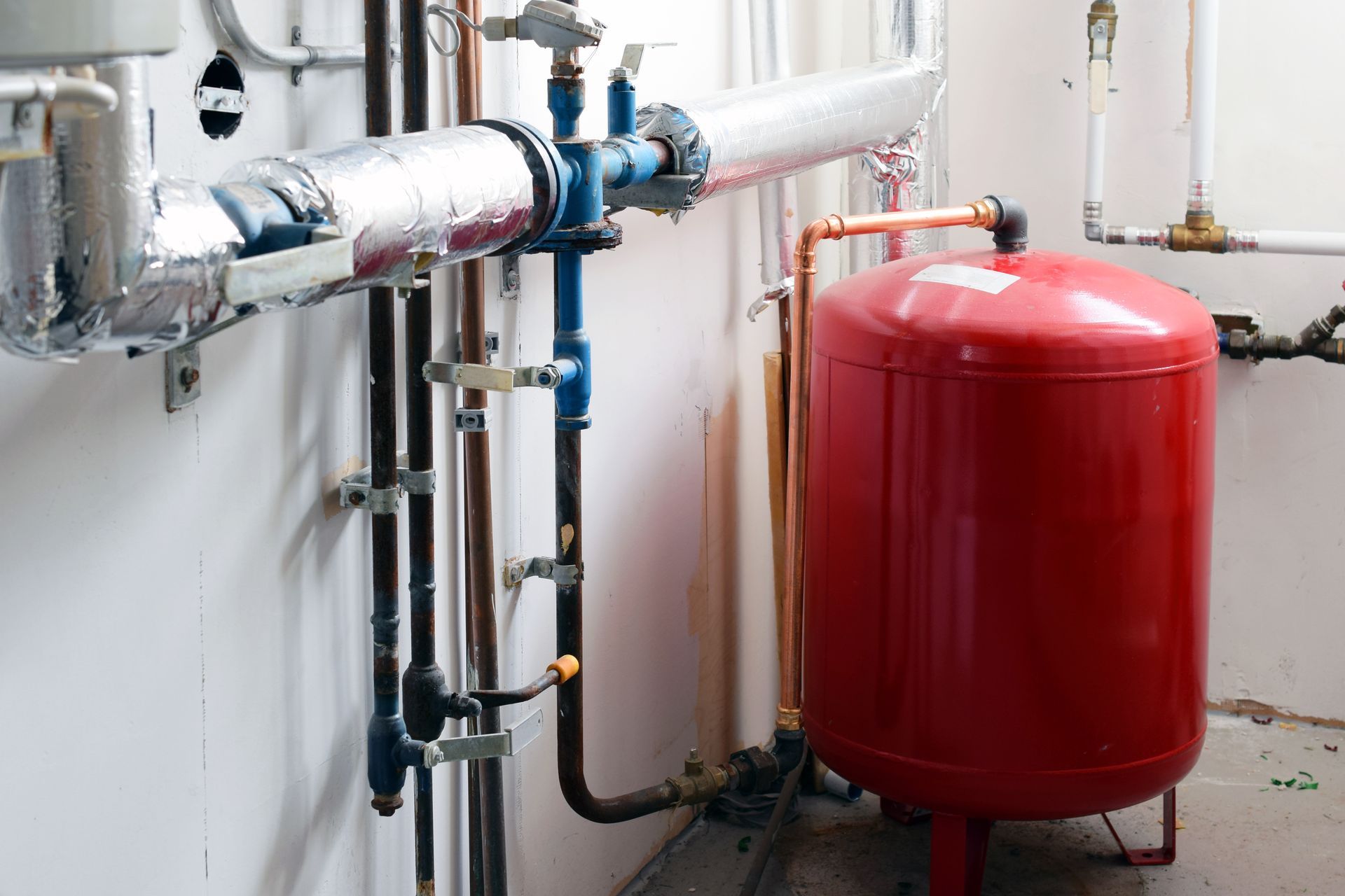 Plumbing system with insulated pipes, valves, and a red expansion tank against a white wall. Plumbing system with insulated pipes, valves, and a red expansion tank against a white wall.