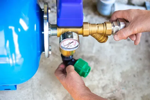 Person working on a blue water tank with brass fittings, a gauge, and black pipe connections. Person working on a blue water tank with brass fittings, a gauge, and black pipe connections.