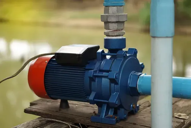 Blue water pump with red motor, gray pipe, and blue pipes on a wooden surface by a pond.