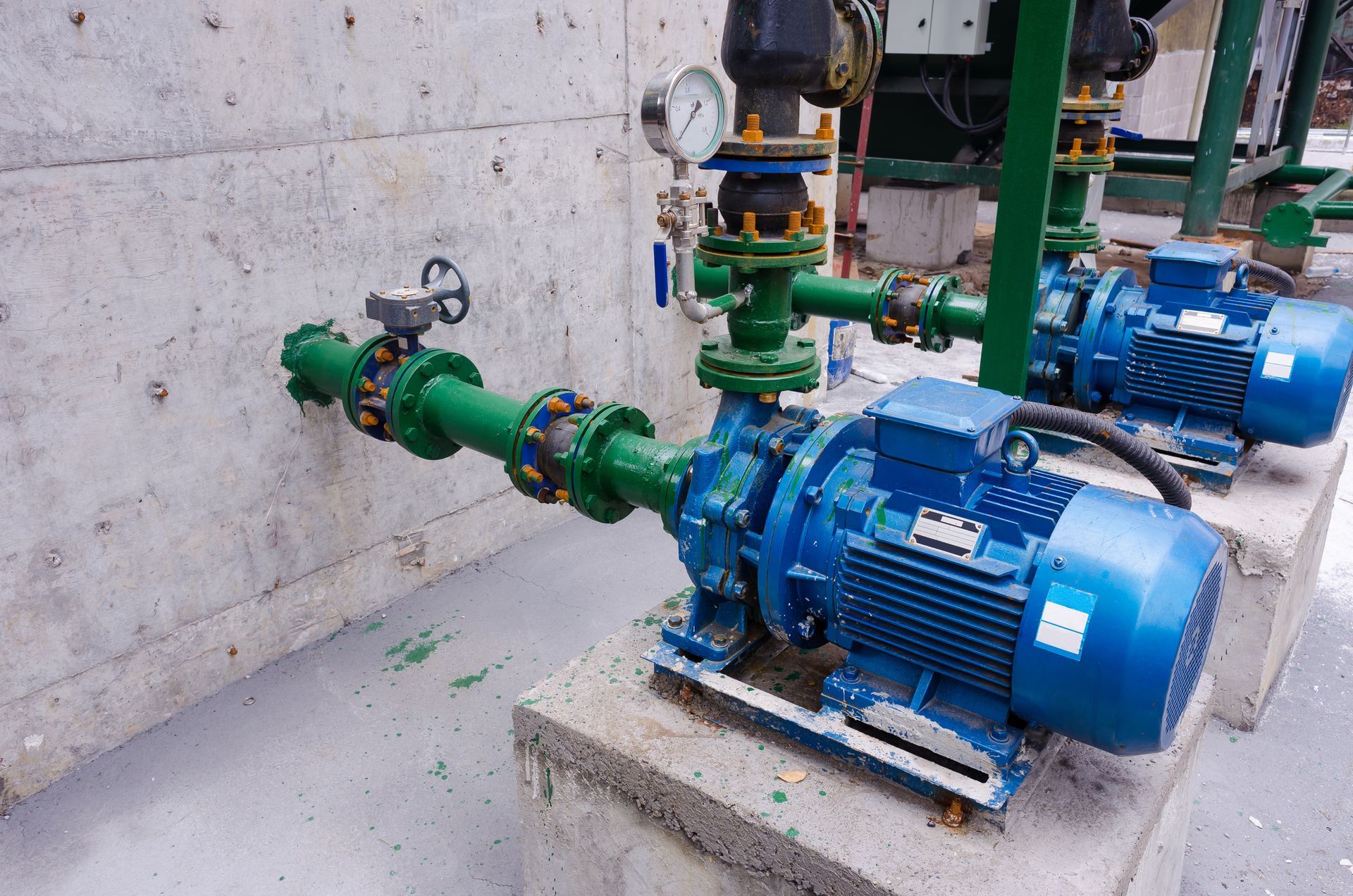 Two blue pumps with green piping attached to a concrete wall in an industrial setting.