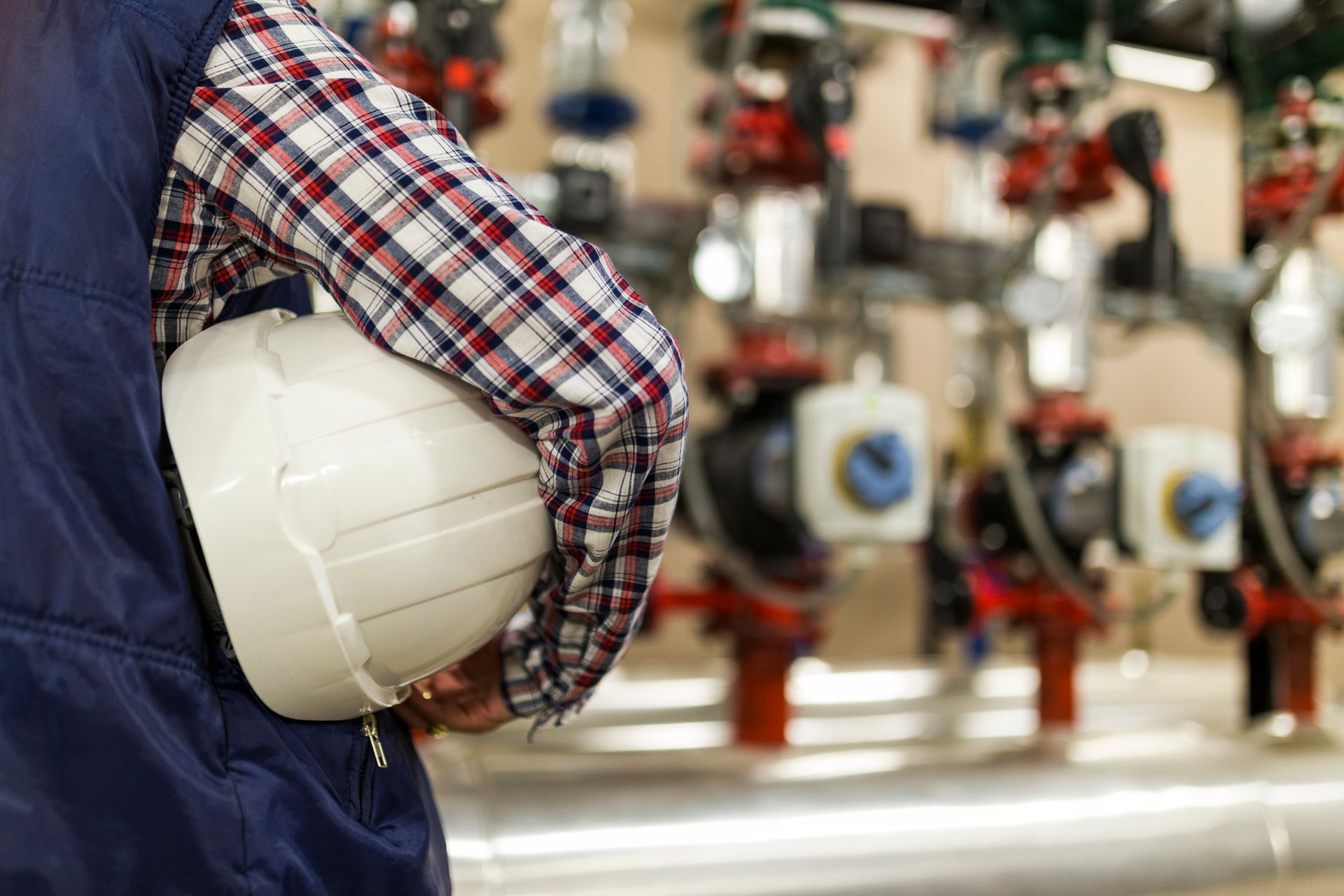 Person in work clothes holding a white hard hat near industrial pipes and machinery.