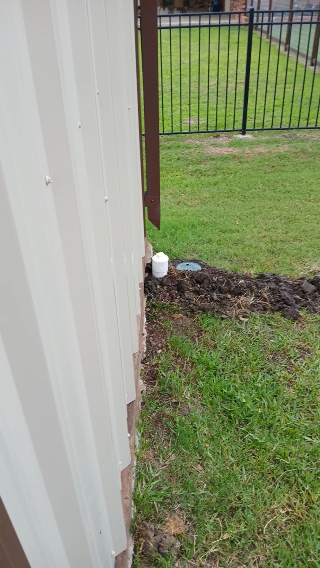 White corrugated metal wall with a white pipe leading into a small dirt patch on green grass.