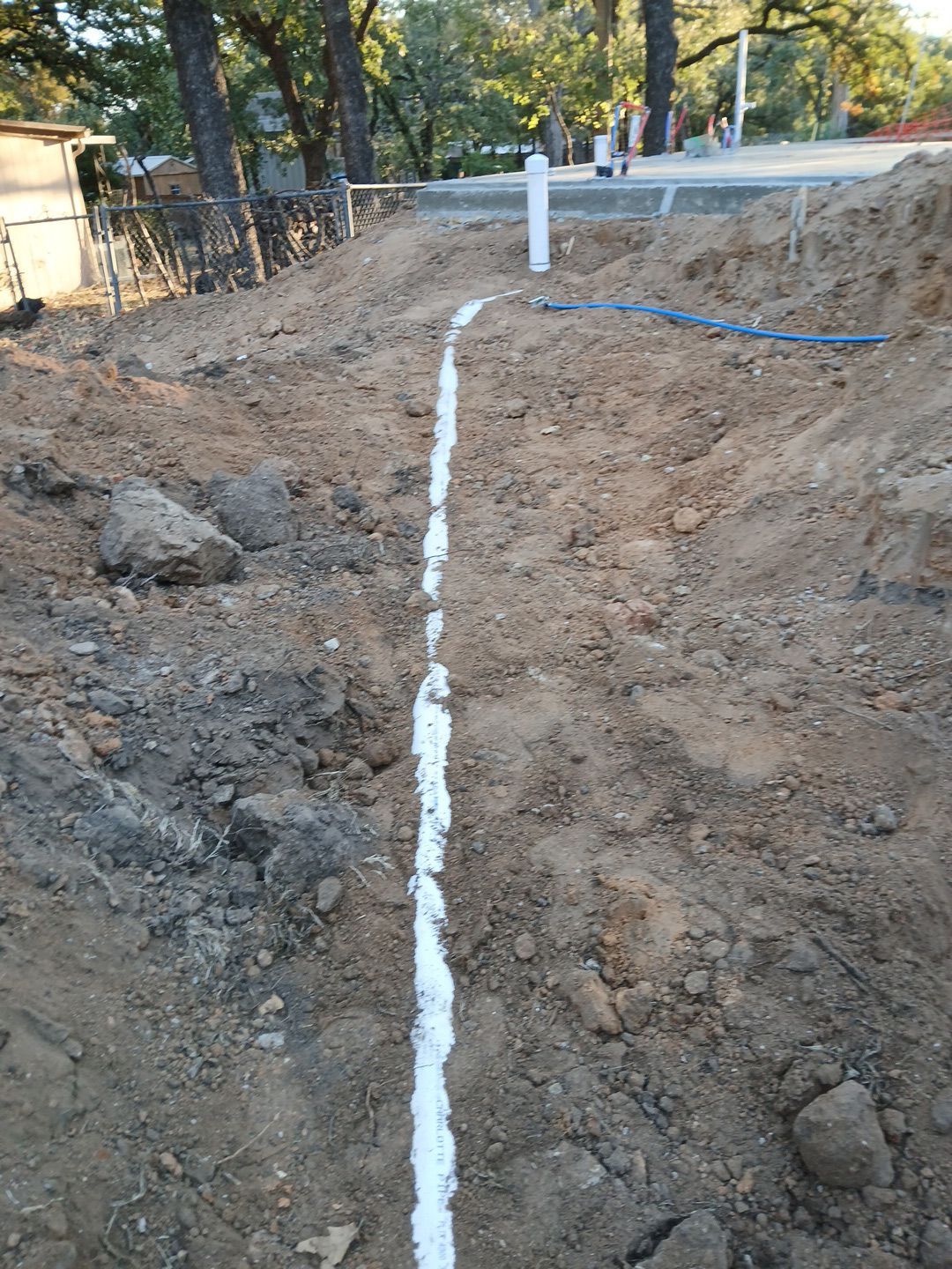 Trench in dirt with white material, possibly a pipe, running lengthwise, with a vertical pipe at the end.
