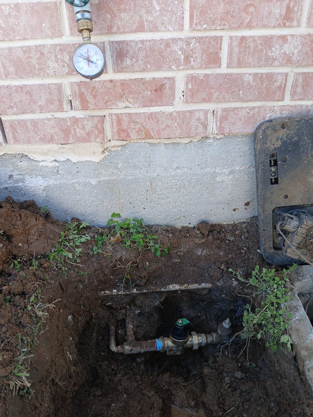 Water meter box in a dug-up area near a brick wall, with a gauge visible above.