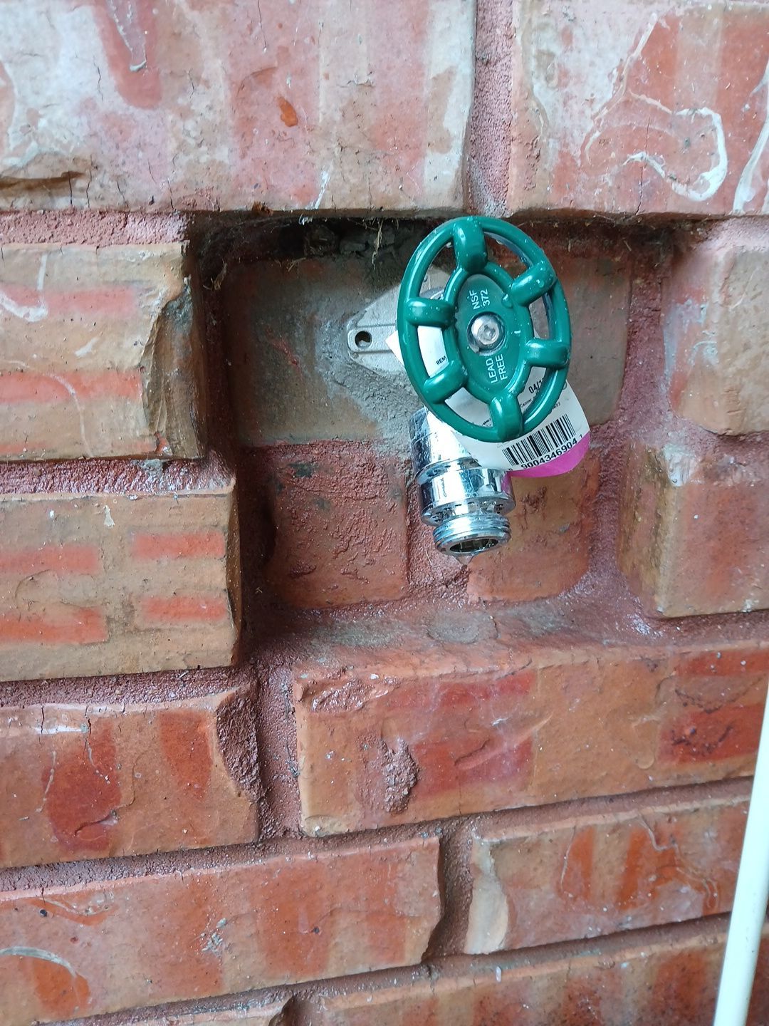 Green-handled outdoor water faucet installed in a red brick wall.