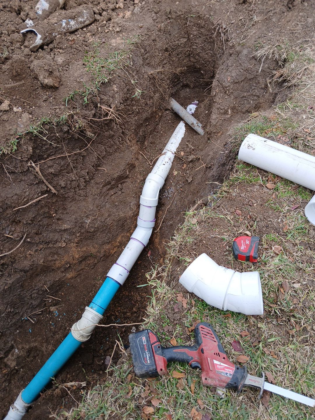 Plumbing repair: buried pipes, elbows, and a saw are in a dirt trench outdoors.