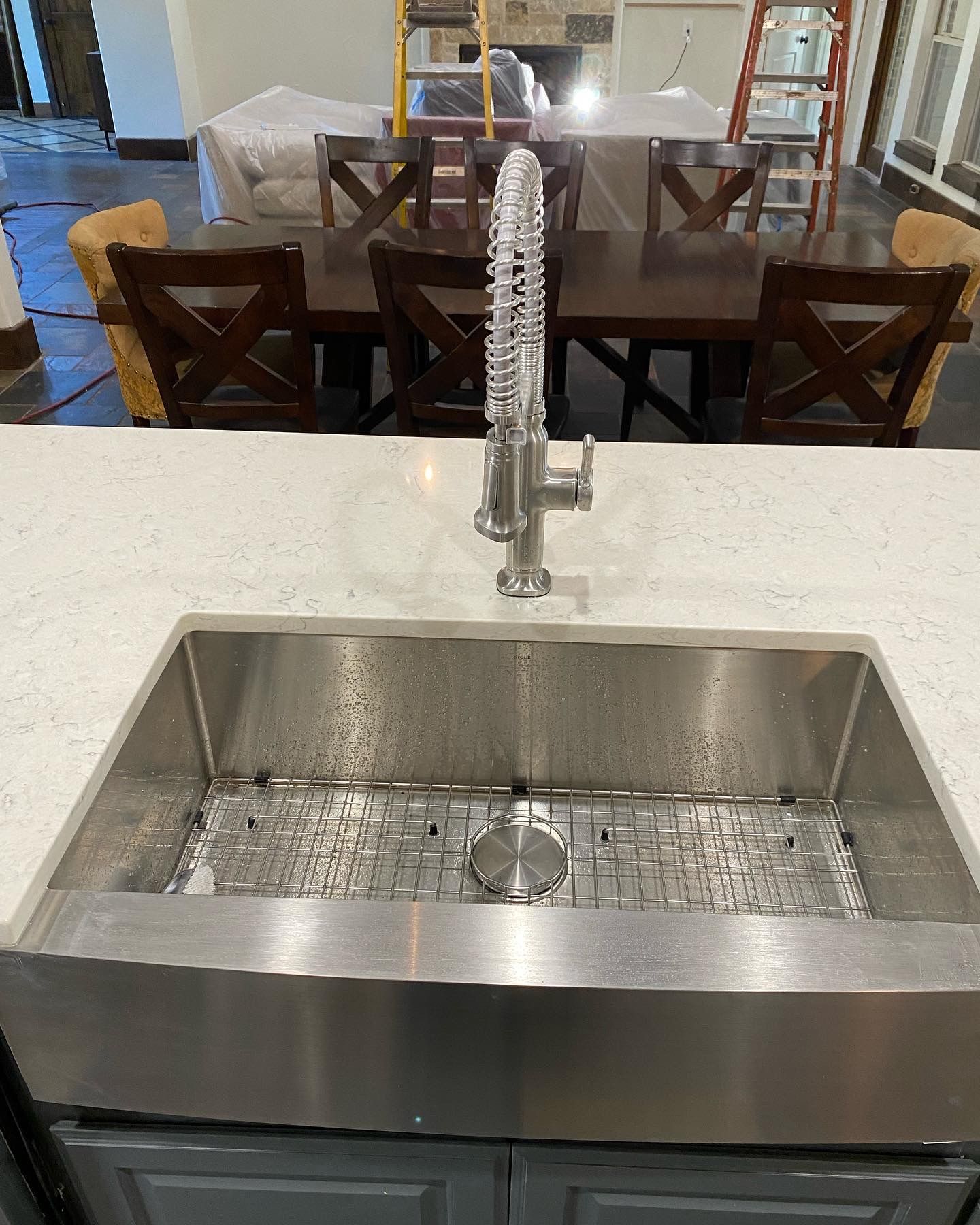 Stainless steel kitchen sink with faucet, white countertop, and dark wood dining table in background.
