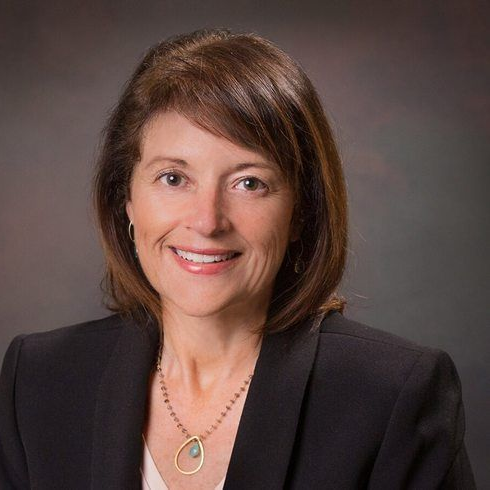 Professional headshot of a smiling woman in a black blazer and necklace against a gray background