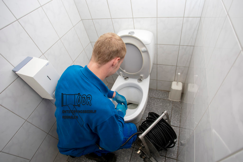 Een servicemonteur in een blauw uniform inspecteert een toilet met een rioolcamera in een betegelde badkamer.