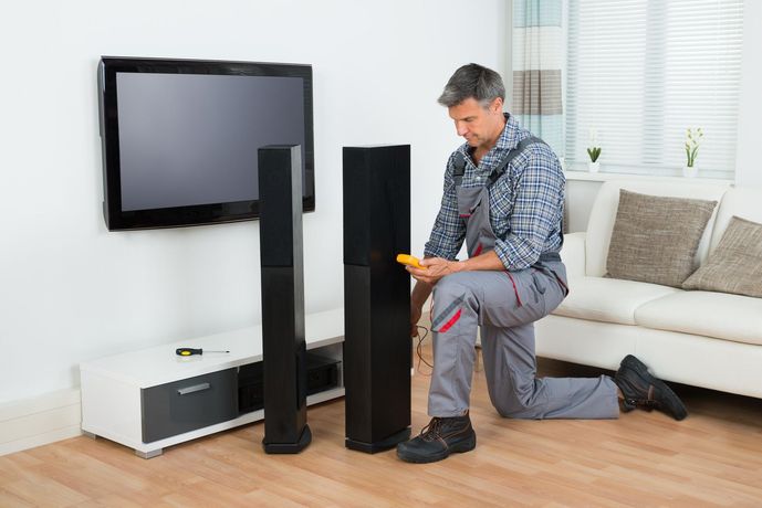 Man assembling a tall black speaker beside a wall-mounted TV in a bright living room.