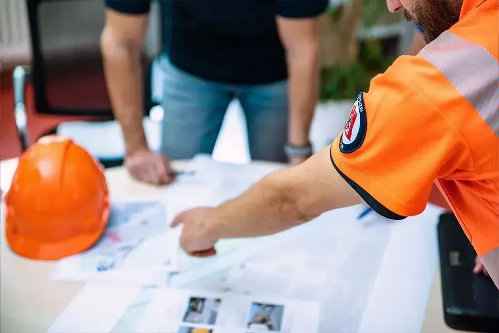 Ein Mann in einem orangefarbenen Hemd sitzt an einem Tisch und betrachtet einen Bauplan.