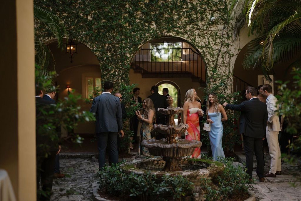 People in formal attire gather around a fountain in a courtyard. 