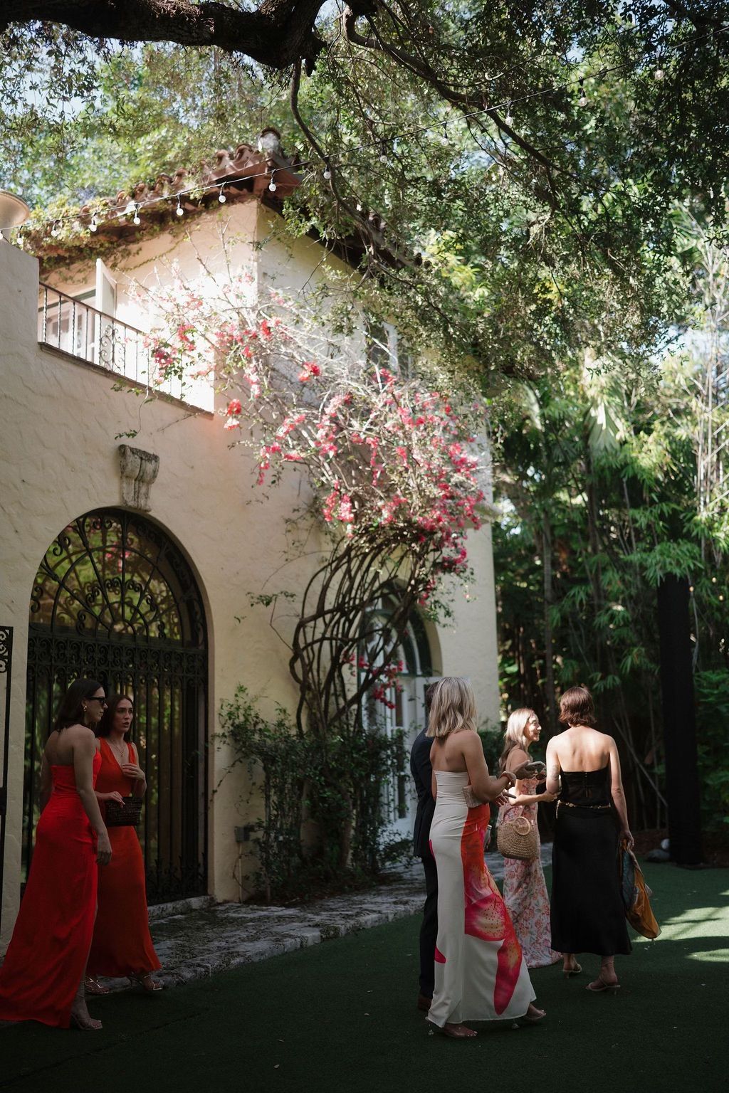 Wedding guests in gowns outside a stucco building with a flowering vine.