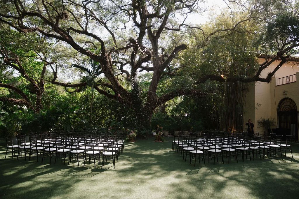 Rows of chairs set up on green lawn for an outdoor event, beneath large trees, next to a building.