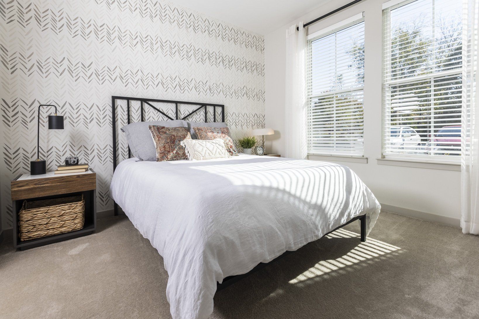 A bedroom with a bed , nightstand , lamp and two windows.
